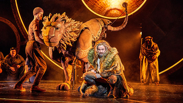 A man on stage wearing a fur coat in front of a life-sized puppet of a lion lit in orange light