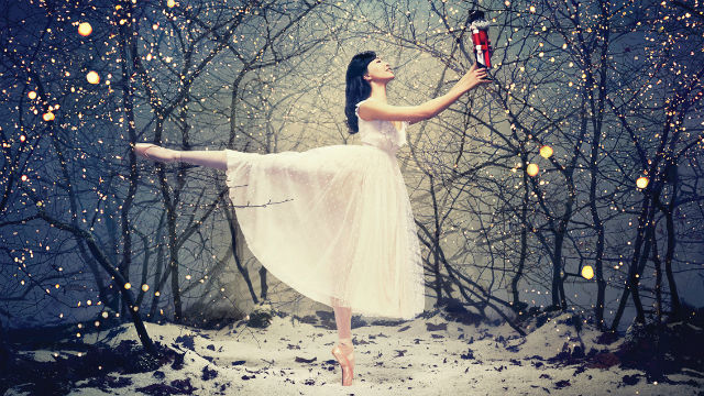 A ballet dancer in a beautiful white dress stands in the middle of a snowy forest holding a nutcracker doll out in front of her.
