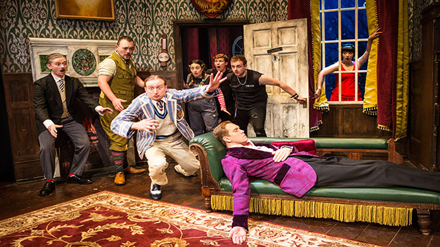 The cast of The Play That Goes Wrong look startled and ready to jump of stage while a man lies unconscious on a couch.