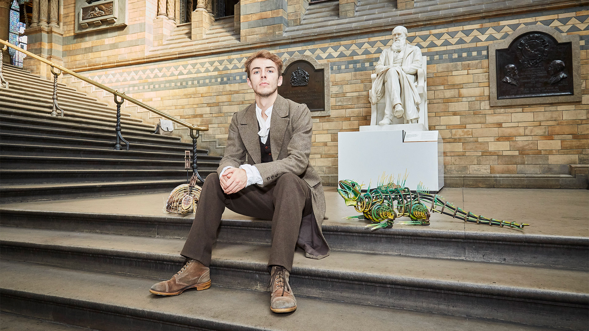 Bradley Foster poses on the steps of the Natural History Museum beside a small dinosaur model.