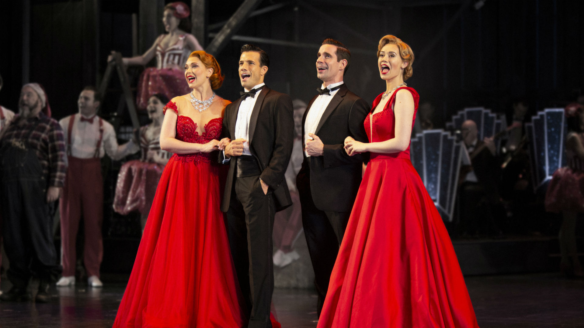 Irving Berlin’s White Christmas at The Dominion Theatre. Credits: Catherine Ashmore and Ellie Kurttz. Image courtesy of Raw PR. Two men in tuxedos stand arm-in-arm with two girls in red ballgowns. All four are singing.