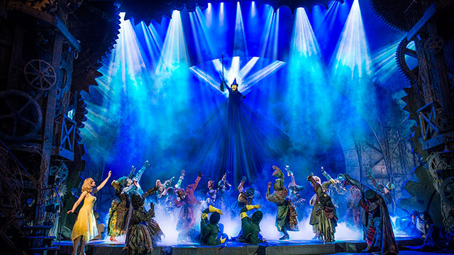 The cast of Wicked the Musical on stage pointing to the witch who is elevated under blue lights