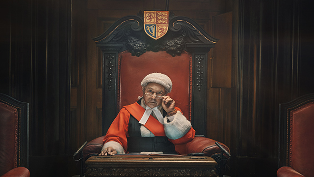 Un juge portant une toge rouge et l'iconique coiffe blanche est assis sur son siège regardant l'audience à travers ses lunettes d'un air sceptique.