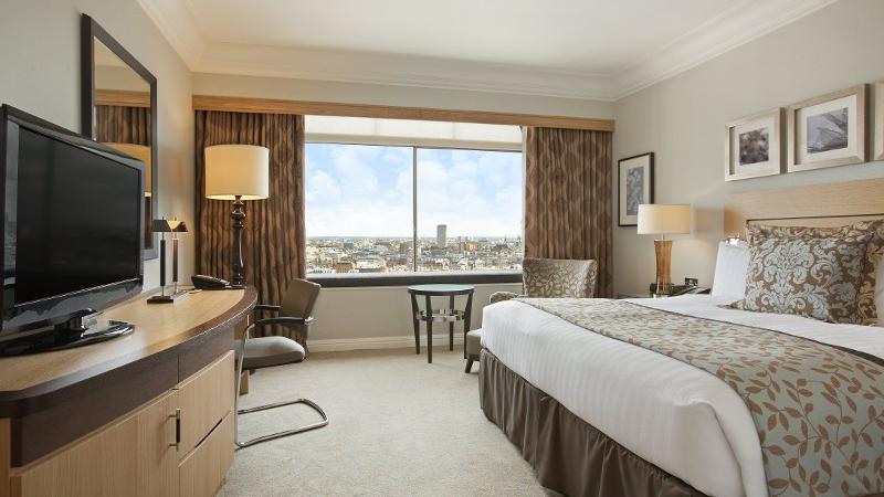 A bedroom at London Hilton on Park Lane, with a large bed, television and seating area, with views in the background of London's skyline from the large window.