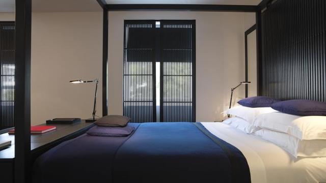 Глядя через двуспальную кровать на окна. Кровать застелена синим пуховым одеялом и белым постельным бельем, с черным изголовьем, сочетающимся с жалюзи на окне.