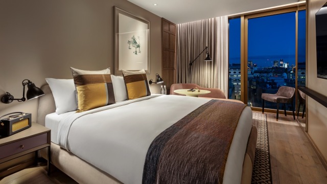 A room at The Londoner Hotel, with a large bed and views over London at night from a floor-to-ceiling window.