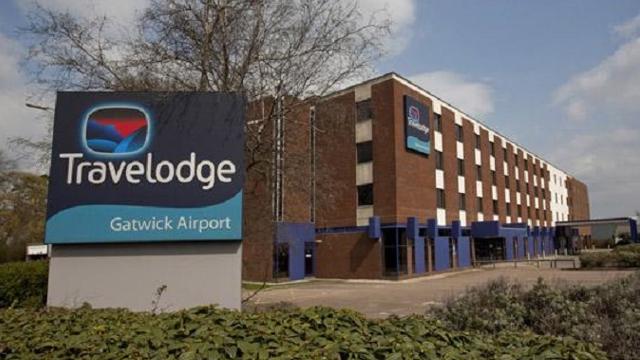 Вывеска Travelodge с логотипом Travelodge и надписью «Travelodge Gatwick Airport» на фоне кирпичного отеля.