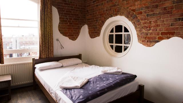 Double bed in hostel room with brick wall, a window overlooking a street and wooden floors.