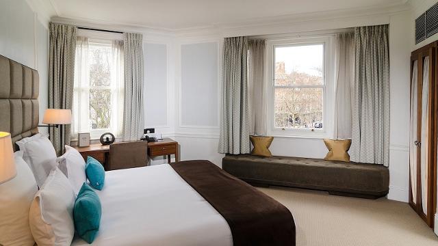 Sloane Square Hotel room featuring white and brown bed linens, colourful blue and gold cushions, and grey curtains.